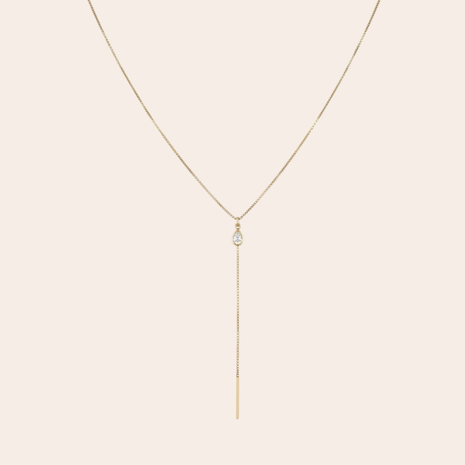 A delicate gold Y-necklace with a teardrop-shaped clear gemstone pendant and a long drop chain, displayed against a solid pale pink background.