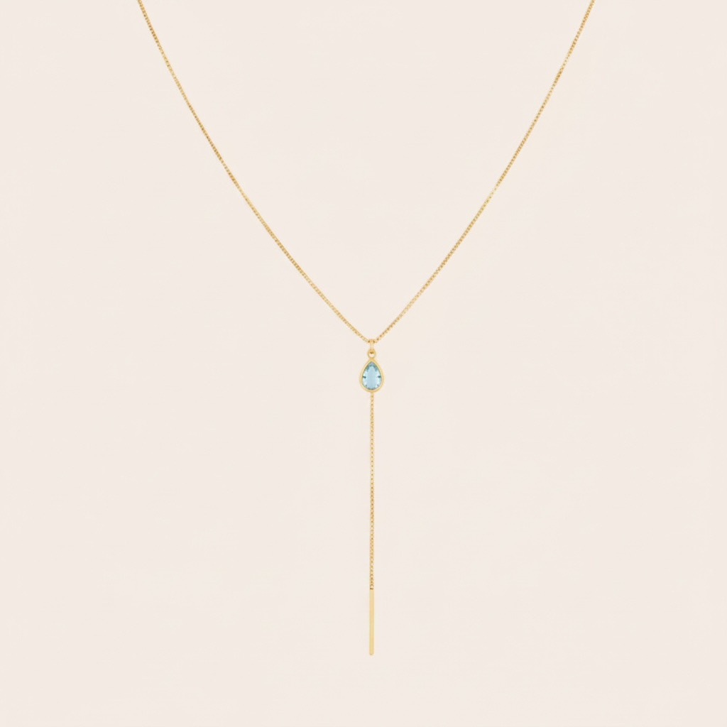 A delicate gold Y-necklace featuring a small light blue teardrop gemstone and a long drop chain, displayed against a solid pale pink background.