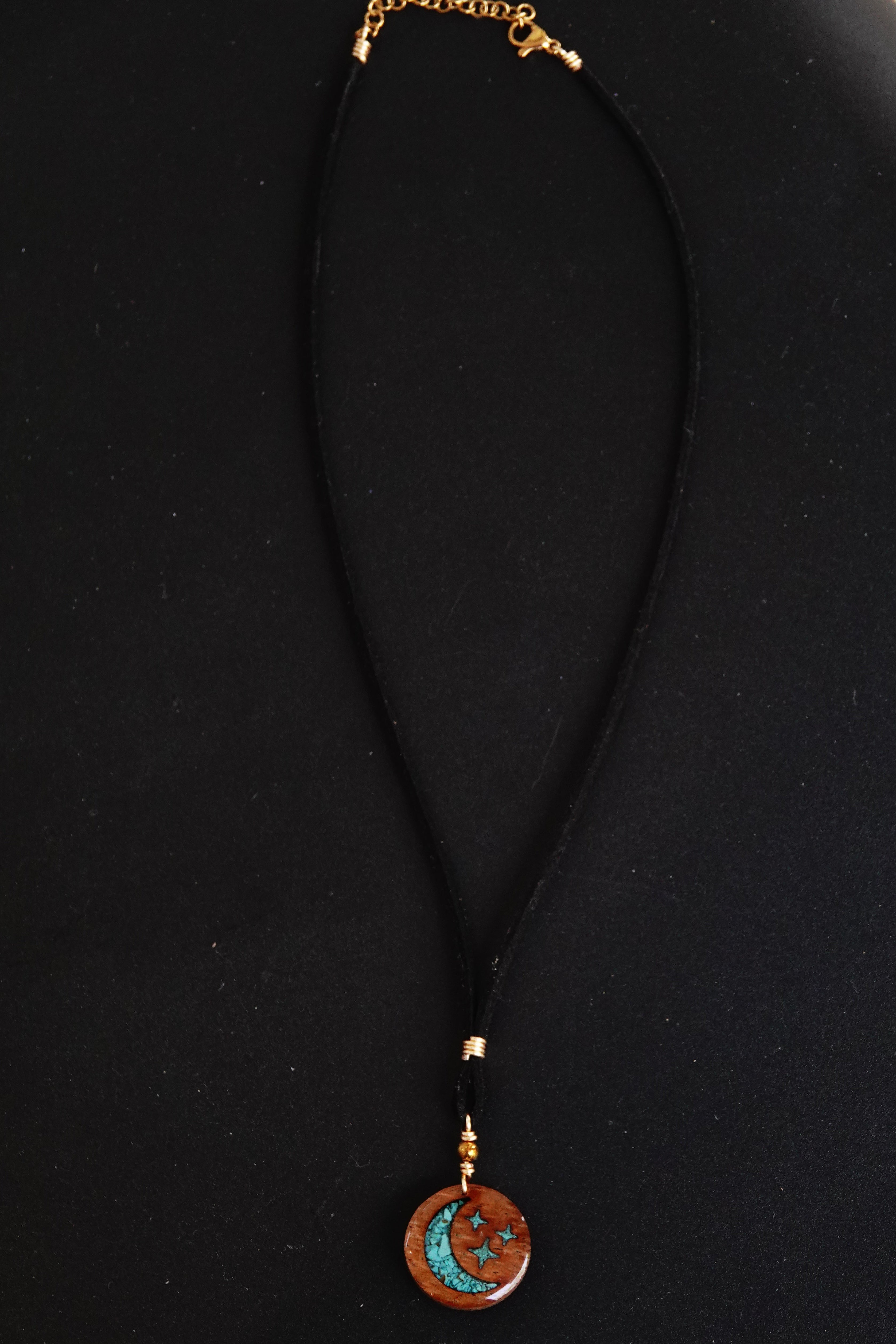 A black cord necklace featuring a circular wooden pendant with a turquoise crescent moon design and gold-toned metal accents, displayed on a black background.