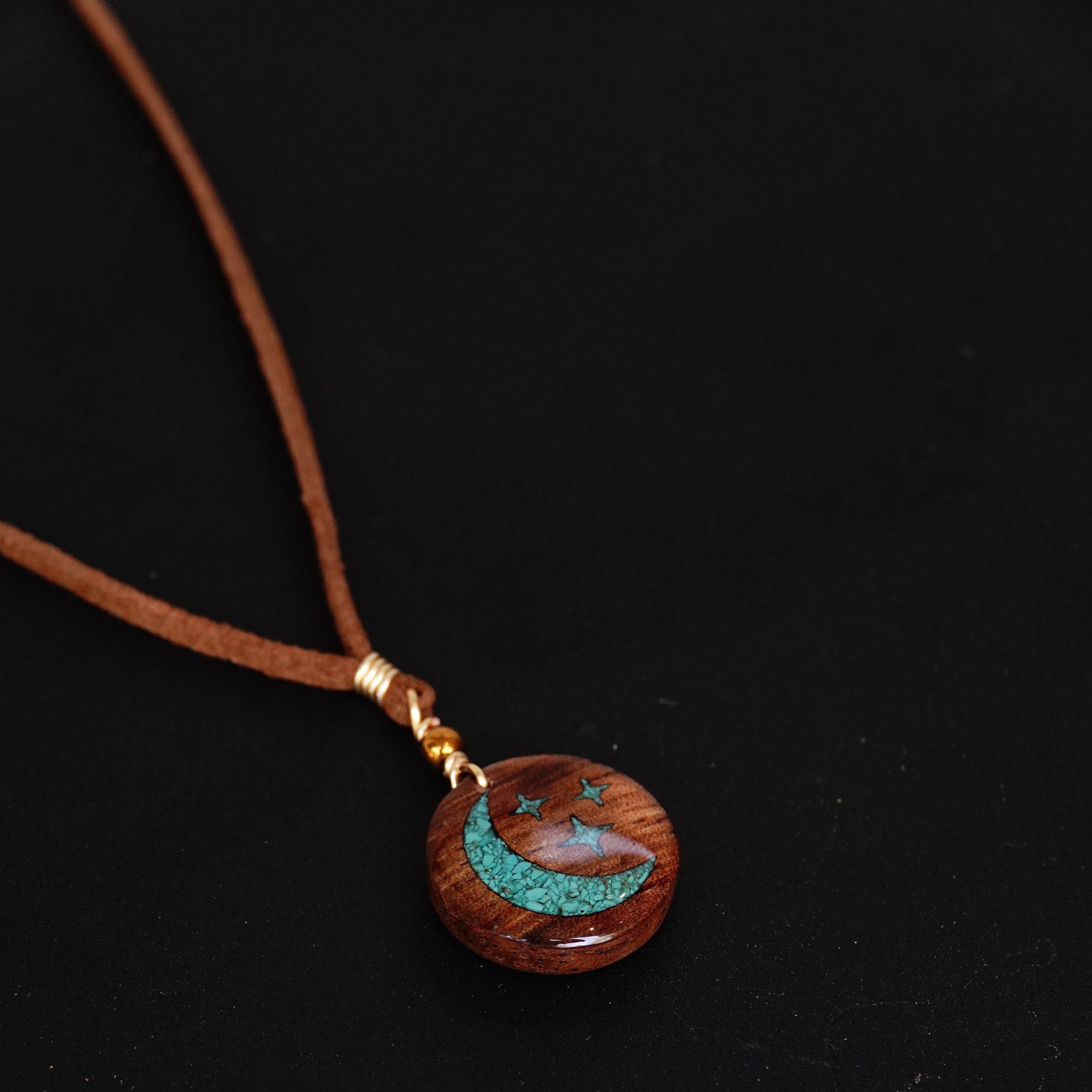 A close-up of a necklace with a round wooden pendant featuring a turquoise crescent moon design and small star details, suspended on a brown cord against a black background.