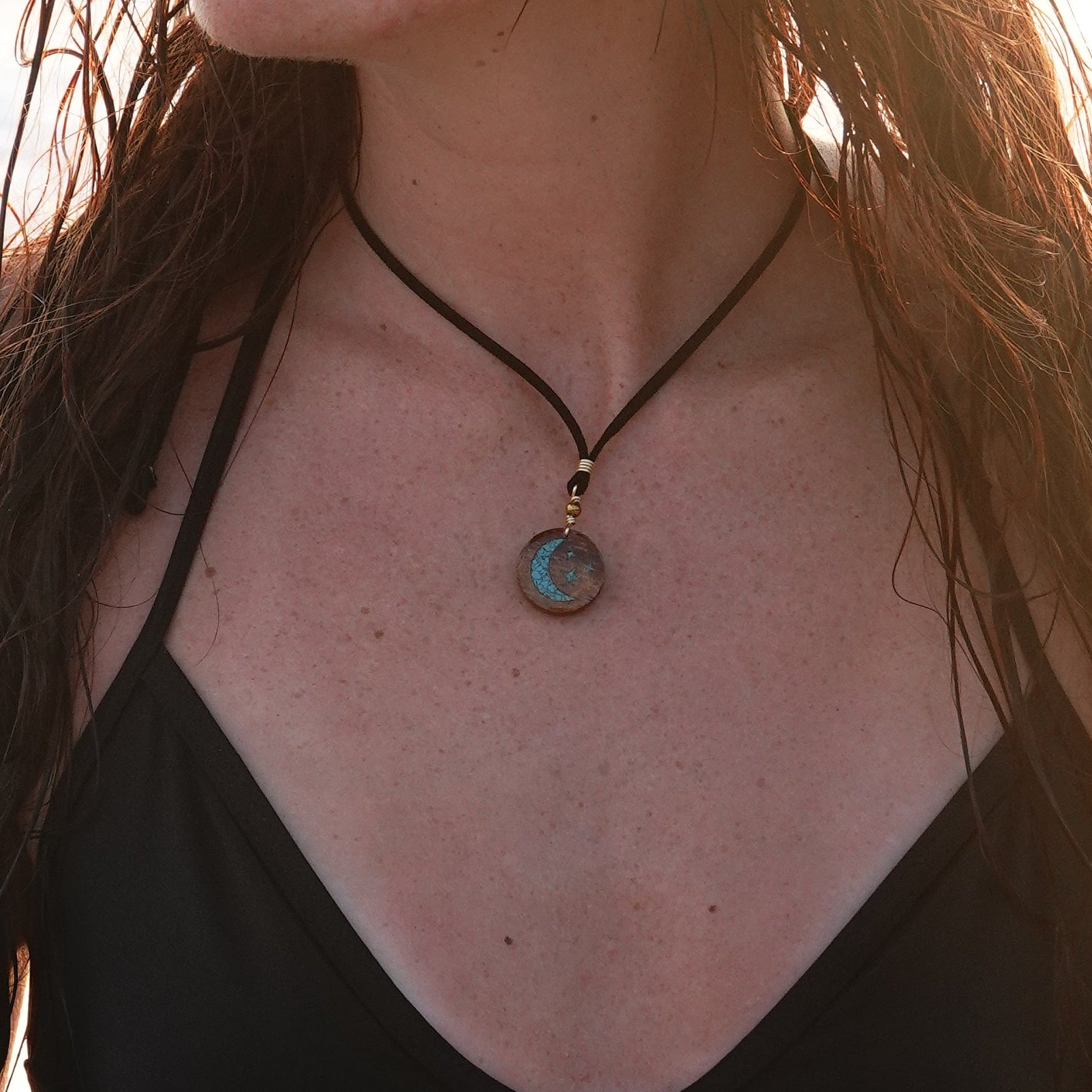 A woman with long, wet brown hair stands on a sandy beach at sunset wearing a black triangle bikini top and a black necklace with a round pendant.