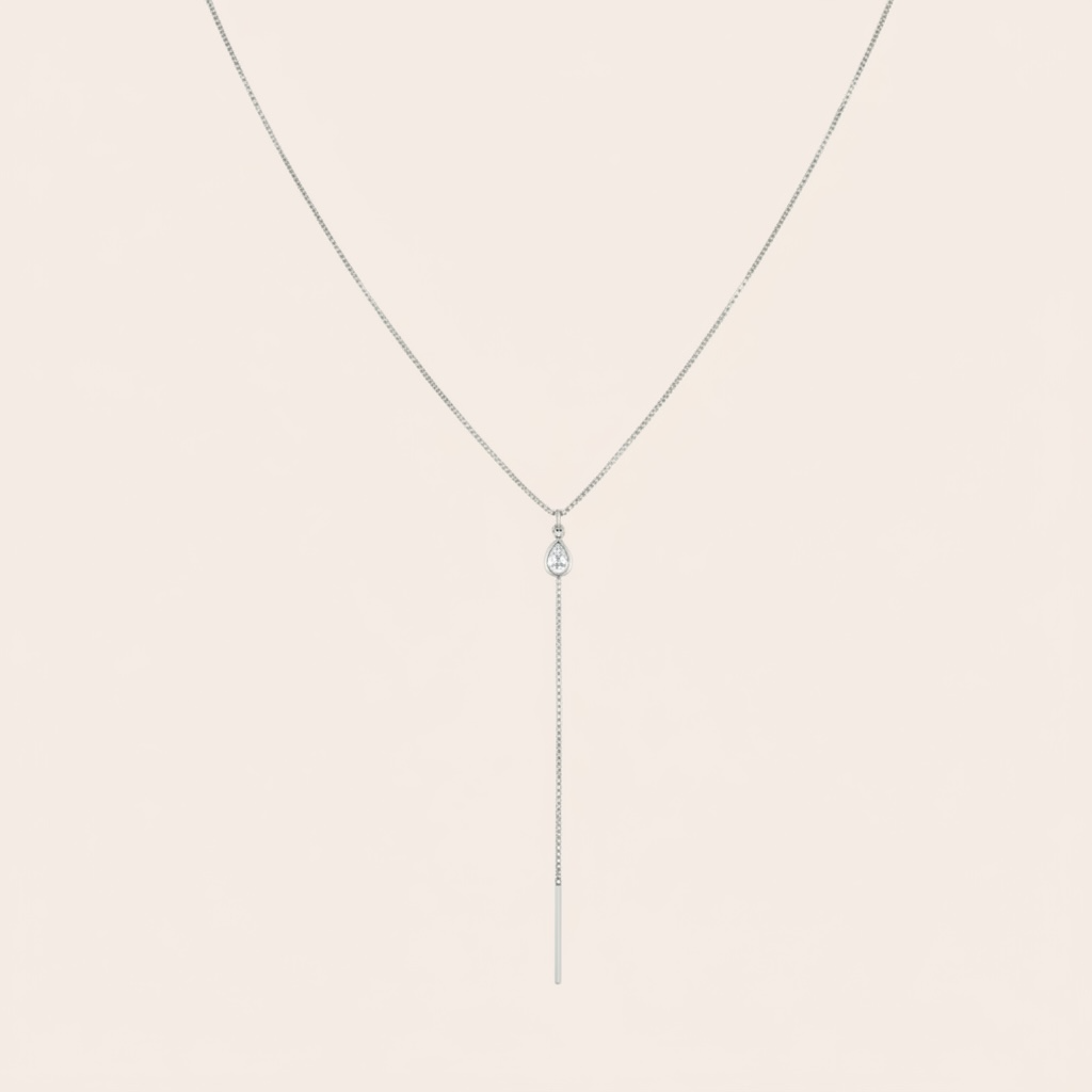 A delicate silver-toned lariat necklace with a teardrop-shaped clear gemstone and a long vertical bar pendant, displayed against a solid pale beige background.