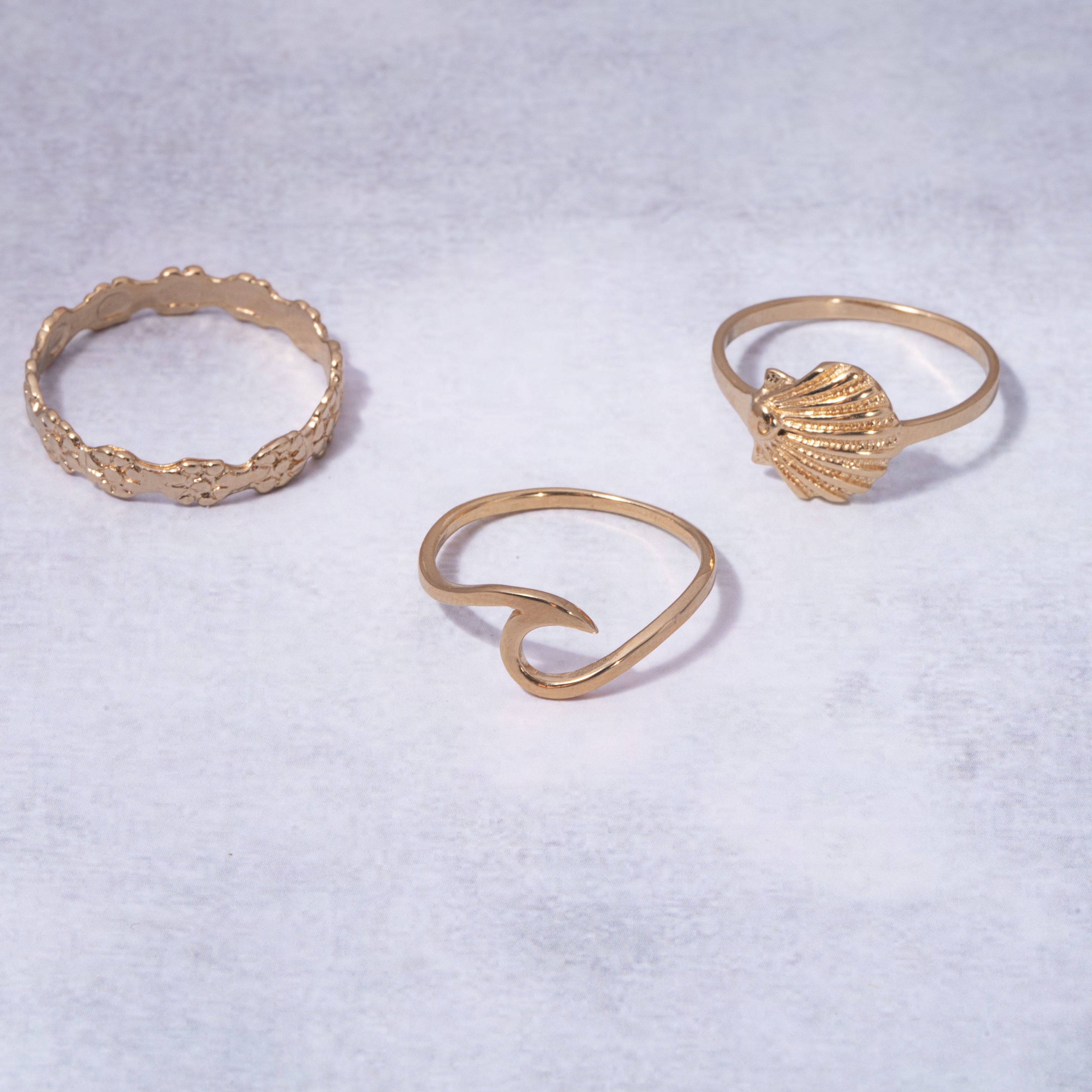 Three gold rings displayed on a textured light gray background: a textured band ring, a heart-shaped ring, and a textured dome ring.