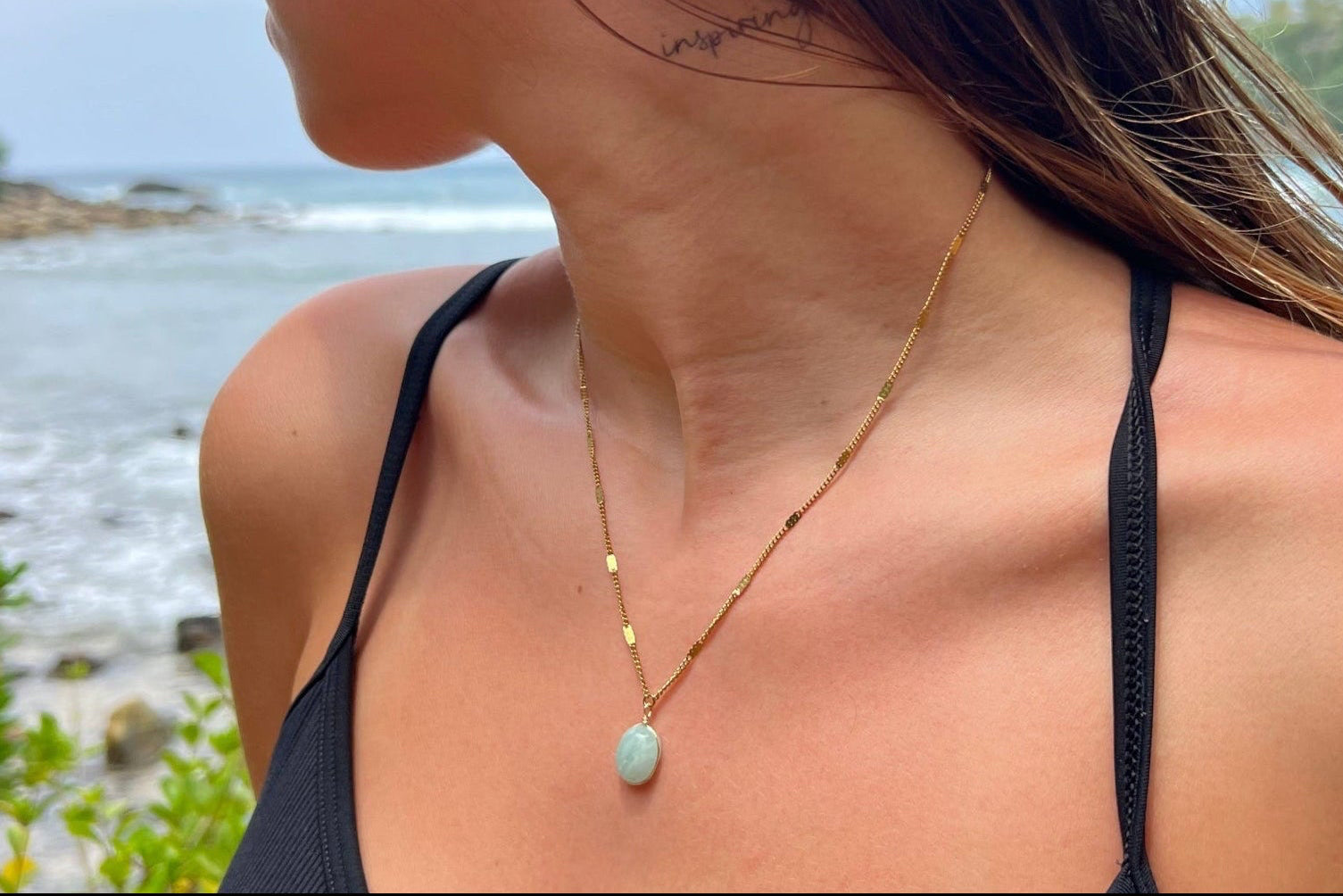 Woman wearing a black tank top with a blue stone necklace and earrings, standing by a body of water.